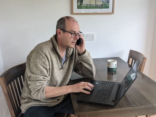 Picture of Steve sitting at table, talking on his mobile phone and typing into his laptop. 